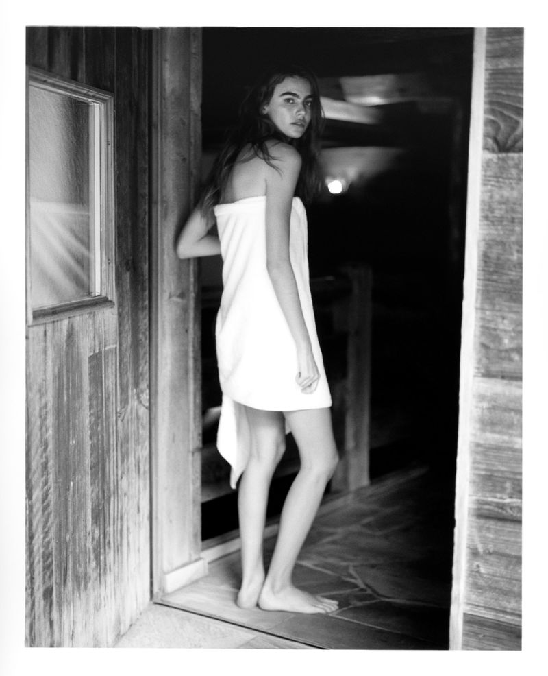 Woman wrapped in a white towel stands in the doorway of a wooden cabin, looking over her shoulder in a black and white photograph.