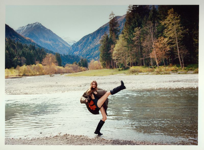 Woman in a puffy coat and boots playfully kicks her leg up in a river with a mountain and forest landscape behind her on an overcast day.