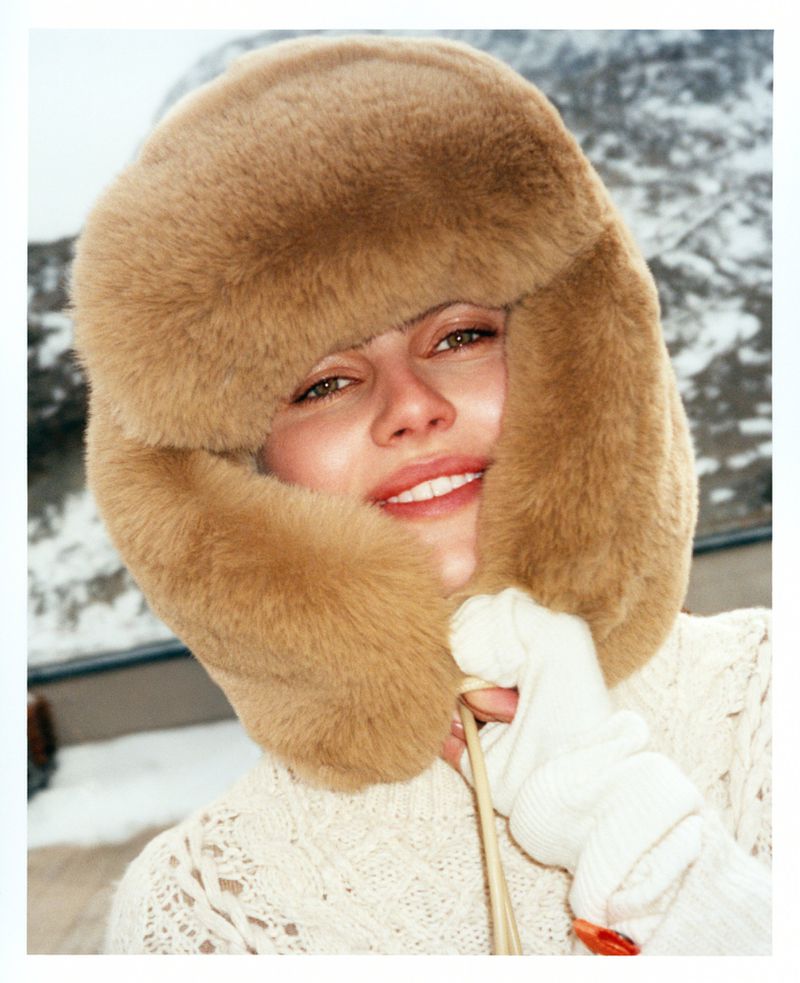 Portrait of smiling woman in fur hat and white gloves holding knitting needles against blurred, snow-covered mountain backdrop.