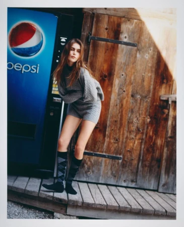 A stylish woman poses in a gray sweater dress and high socks next to a Pepsi vending machine and a weathered wood door in daylight.