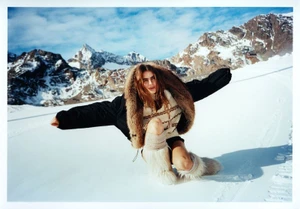 Mia Armstrong crouching on a snowy alpine slope wearing a black jacket with a large fur hood and furry winter boots.