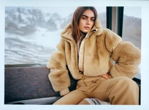 Model Mia Armstrong wearing a camel faux fur jacket and beige trousers inside a cable car overlooking snowy alpine mountains.