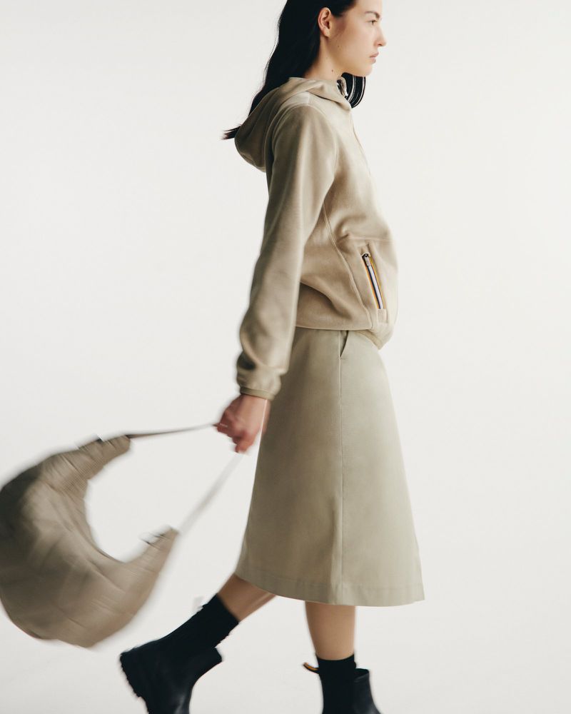 Fashion model wearing a beige hooded sweatshirt and midi skirt walks against a white backdrop holding a beige bag. Black ankle boots complete the look.
