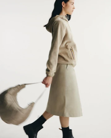 Fashion model wearing a beige hooded sweatshirt and midi skirt walks against a white backdrop holding a beige bag. Black ankle boots complete the look.