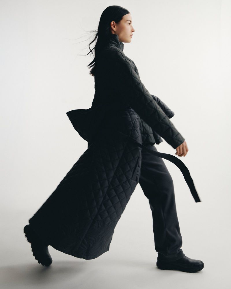 A woman in a quilted coat strides against a white backdrop in a full shot, showcasing fashion in motion with natural light.