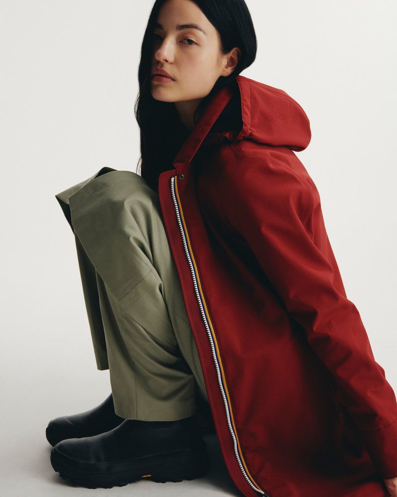 Brunette woman in a hooded raincoat and green trousers sits in a studio, looking at the camera. The photo features an eye-level angle.