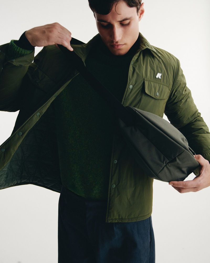 A man opens his olive-green quilted jacket indoors, revealing a dark green sweater and dark trousers, holding a matching bag.