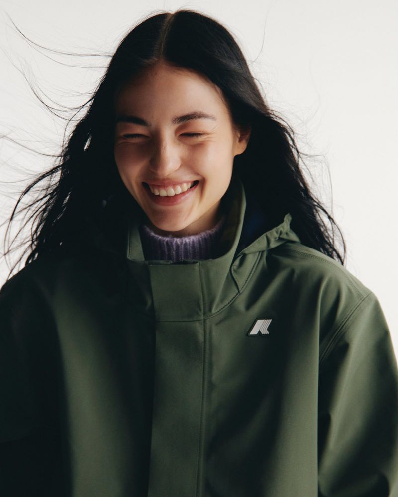 Smiling woman in a green jacket, standing indoors against a white background, showcasing fashion and beauty with a cheerful expression.