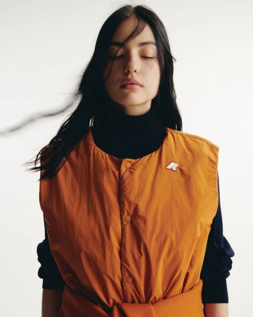 Portrait of a young woman with eyes closed wearing a rust-colored vest over a black turtleneck in a bright studio setting.