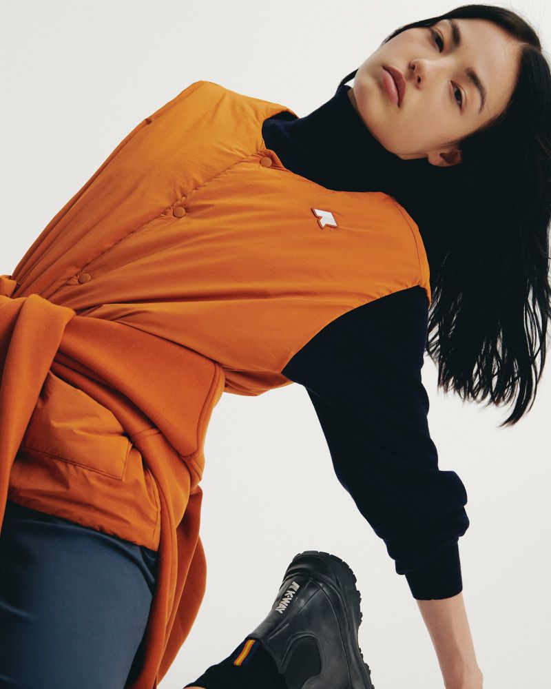 Indoor shot of a young woman in a black turtleneck and orange vest, balancing with one black boot visible in a brightly lit photo.