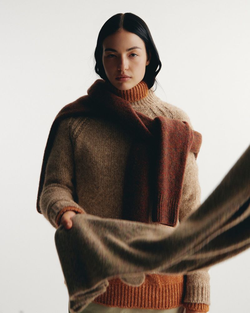 A brunette adult woman in a tan turtleneck sweater and terracotta scarf poses in a portrait on a white backdrop in a studio.