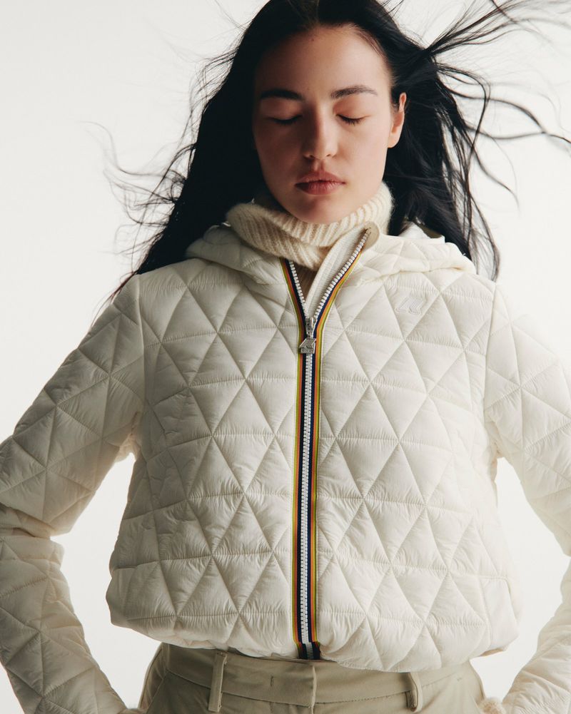 Woman in hooded puffer jacket with closed eyes. Dark hair is blowing around her face. She is wearing a turtleneck sweater.