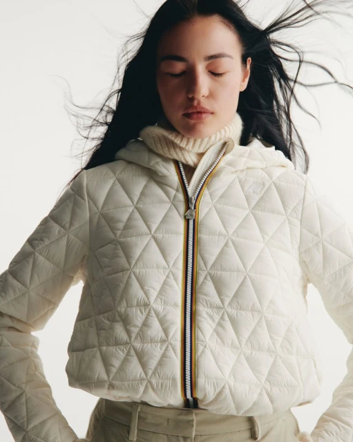 Woman in hooded puffer jacket with closed eyes. Dark hair is blowing around her face. She is wearing a turtleneck sweater.