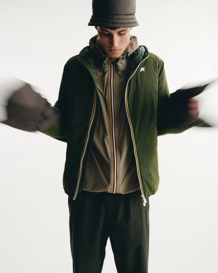 Man wearing a green hooded jacket and bucket hat with motion blur on his hands against a seamless white background.