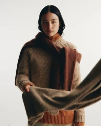 Model with dark hair wearing layered beige and rust-brown knit sweaters against a plain white studio background.