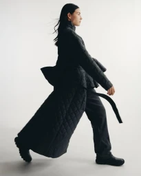 Model walking in profile wearing a long black quilted coat, dark trousers, and black footwear against a clean white backdrop.