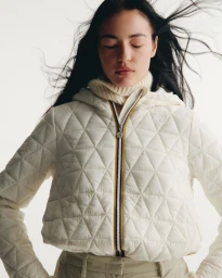 Woman with eyes closed wearing an off-white geometric quilted K-Way zip jacket and knit turtleneck against a white studio backdrop.