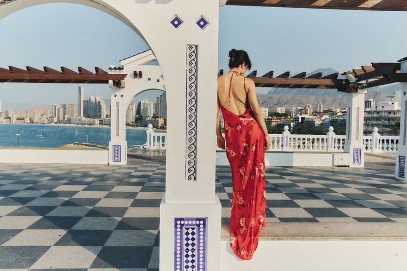 Woman in a red floral dress stands on a checkered terrace overlooking a city and ocean view framed by white architectural elements.