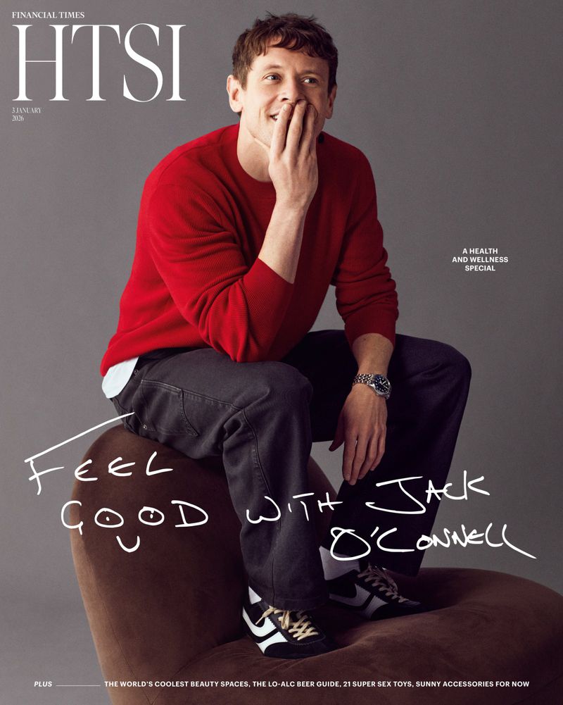 Magazine cover featuring Jack O'Connell wearing red sweater and dark pants while seated on a brown chair against a gray background.