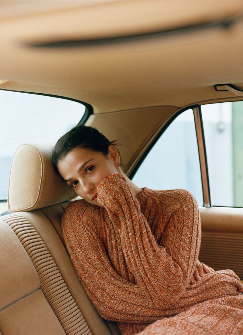A brunette woman in a knit sweater and skirt sits in the back seat of a vintage car, resting her head against the tan leather headrest.