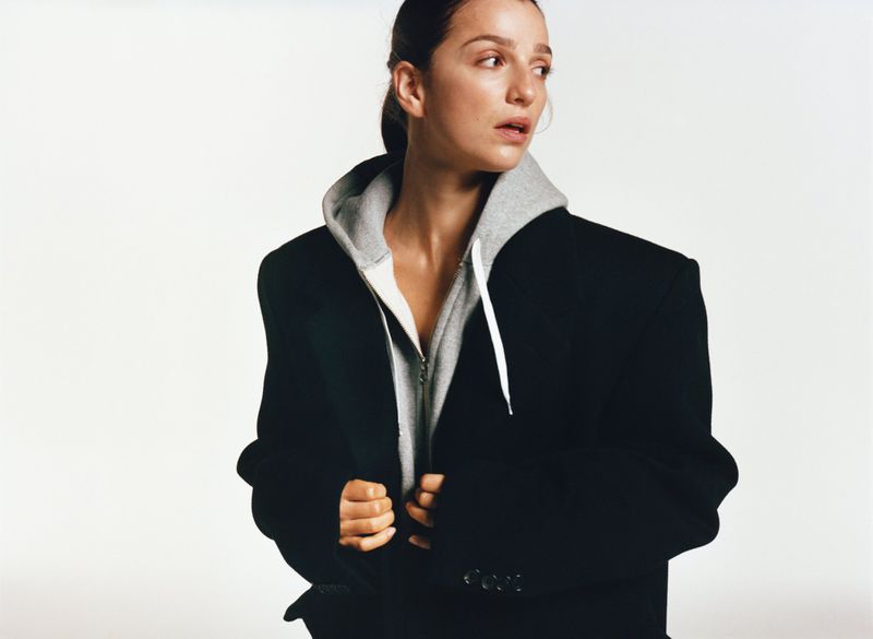 Portrait of a woman wearing a grey hoodie and an oversized black blazer looking away from the camera against a white backdrop.