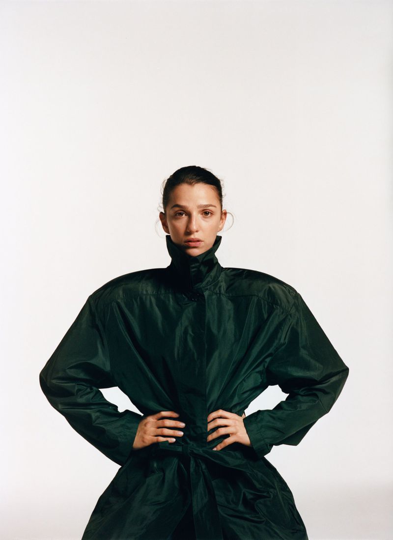 Studio shot of a woman in a dark green, high-collared coat, standing with hands on hips against a white background in portrait orientation.