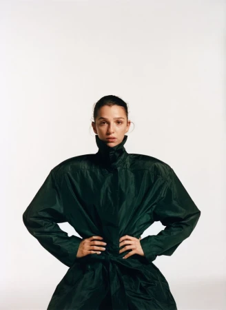 Studio shot of a woman in a dark green, high-collared coat, standing with hands on hips against a white background in portrait orientation.