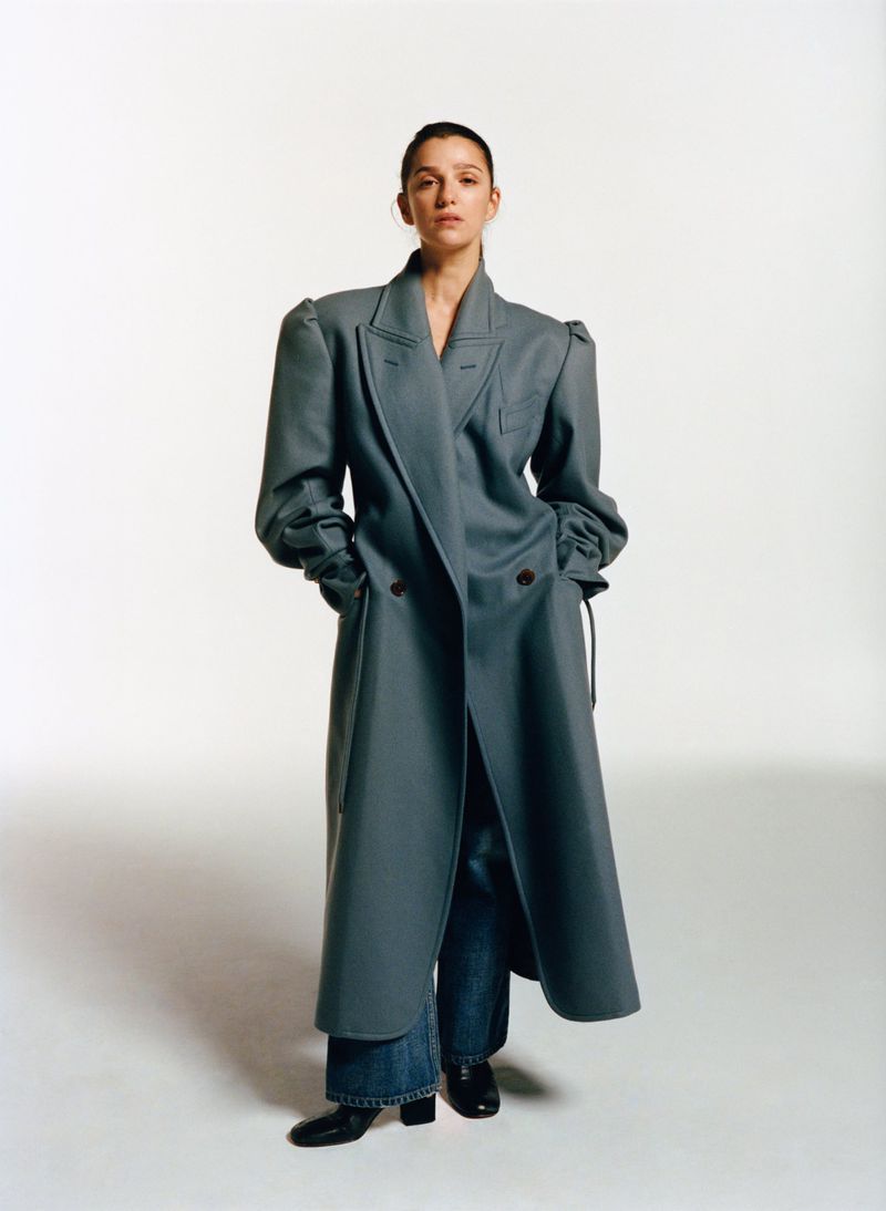 A woman in a long, light blue coat with puffy sleeves and wide-leg jeans poses in a studio setting, showcasing a blend of modern and classic styles.