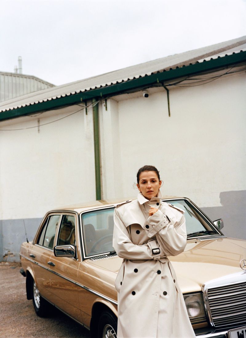 A stylish woman in a beige trench coat stands confidently next to a gold-colored classic Mercedes-Benz car in an urban setting, cloudy day.