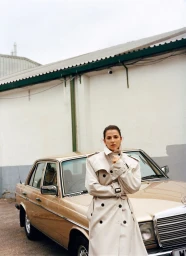 Model wearing an oversized beige trench coat posed outdoors in front of a vintage gold sedan against an industrial building.