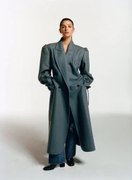 Marisa Abela standing in an oversized slate grey coat with structured shoulders, wide-leg jeans, and black boots against a white backdrop.