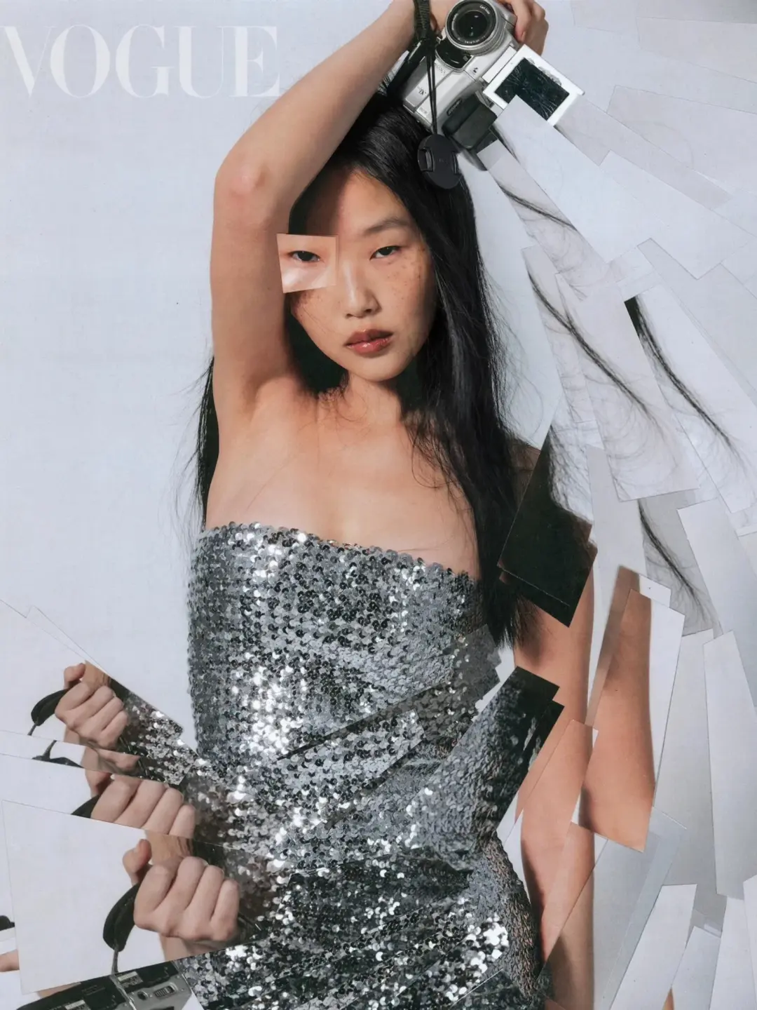 Fashion editorial shot of a person in a silver sequin dress holding a camera, surrounded by photo prints in a photo studio.