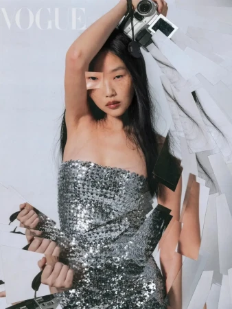 Fashion editorial shot of a person in a silver sequin dress holding a camera, surrounded by photo prints in a photo studio.
