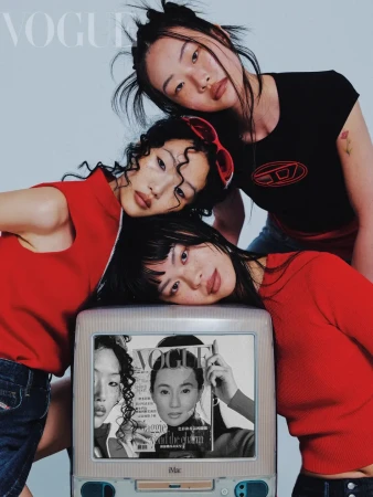 Three young Asian women pose indoors wearing red and black tops, leaning on an old computer monitor displaying a fashion magazine cover.