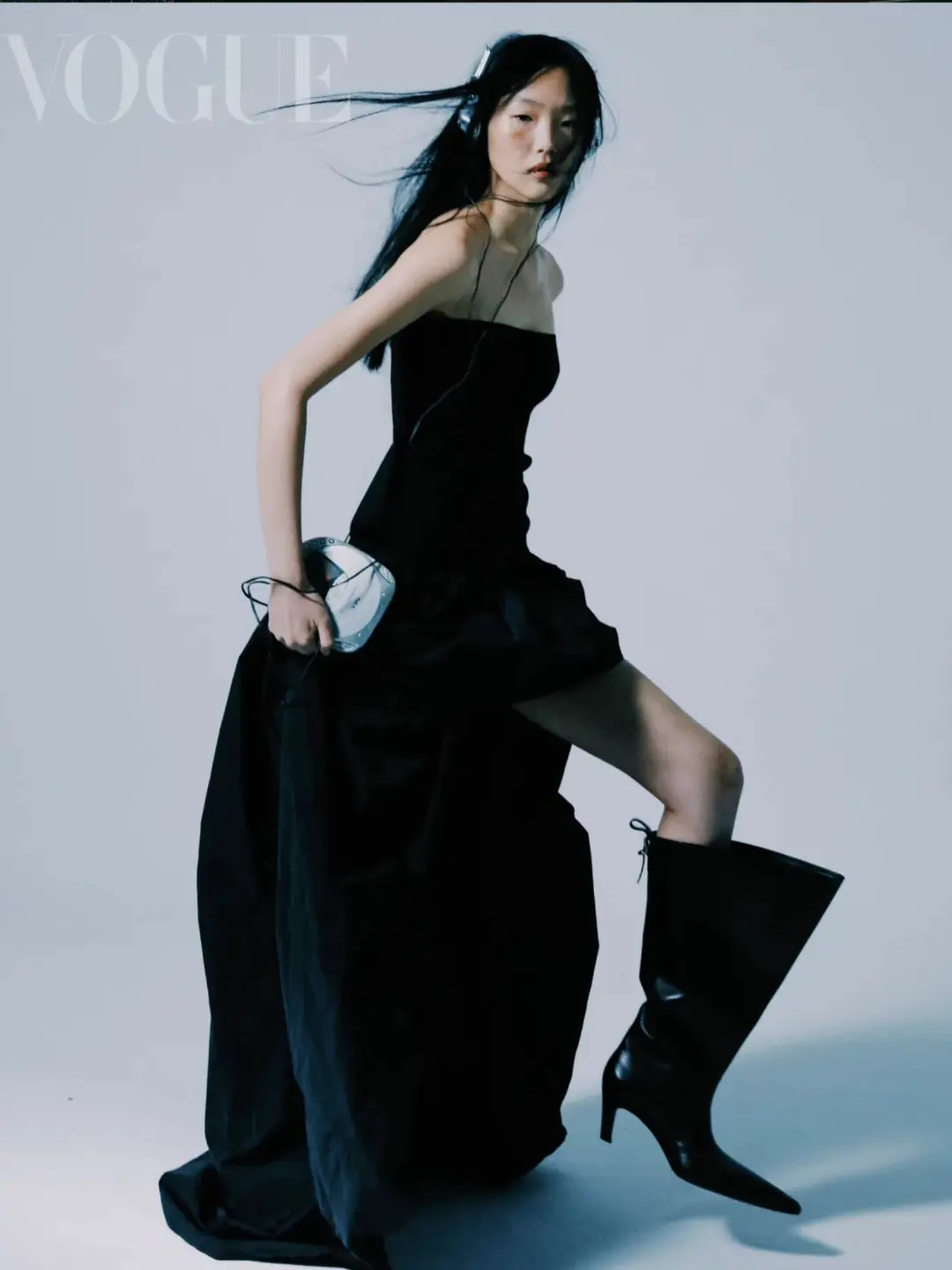 Asian woman in a black dress and boots poses with a disc player, wearing headphones, in a fashion editorial shoot for Vogue magazine.