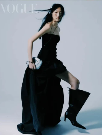 Asian woman in a black dress and boots poses with a disc player, wearing headphones, in a fashion editorial shoot for Vogue magazine.