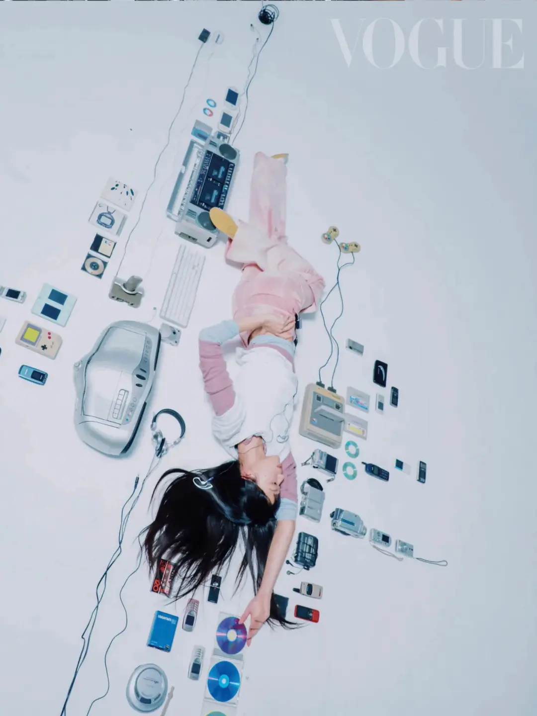 Overhead shot of a young woman lying on a white surface surrounded by vintage electronics, including a cassette player and CDs, with Vogue logo.