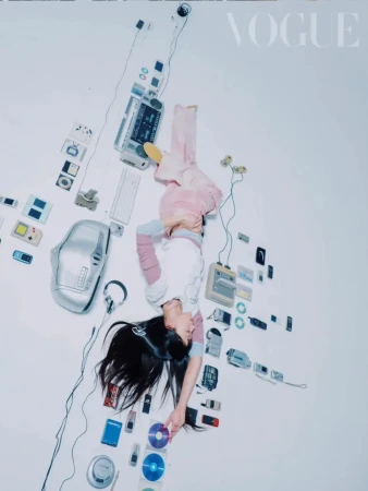 Overhead shot of a young woman lying on a white surface surrounded by vintage electronics, including a cassette player and CDs, with Vogue logo.