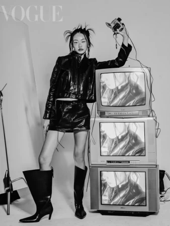 Black-and-white fashion shot of an Asian model in leather outfit, holding a camcorder, standing near stacked retro televisions in a studio.
