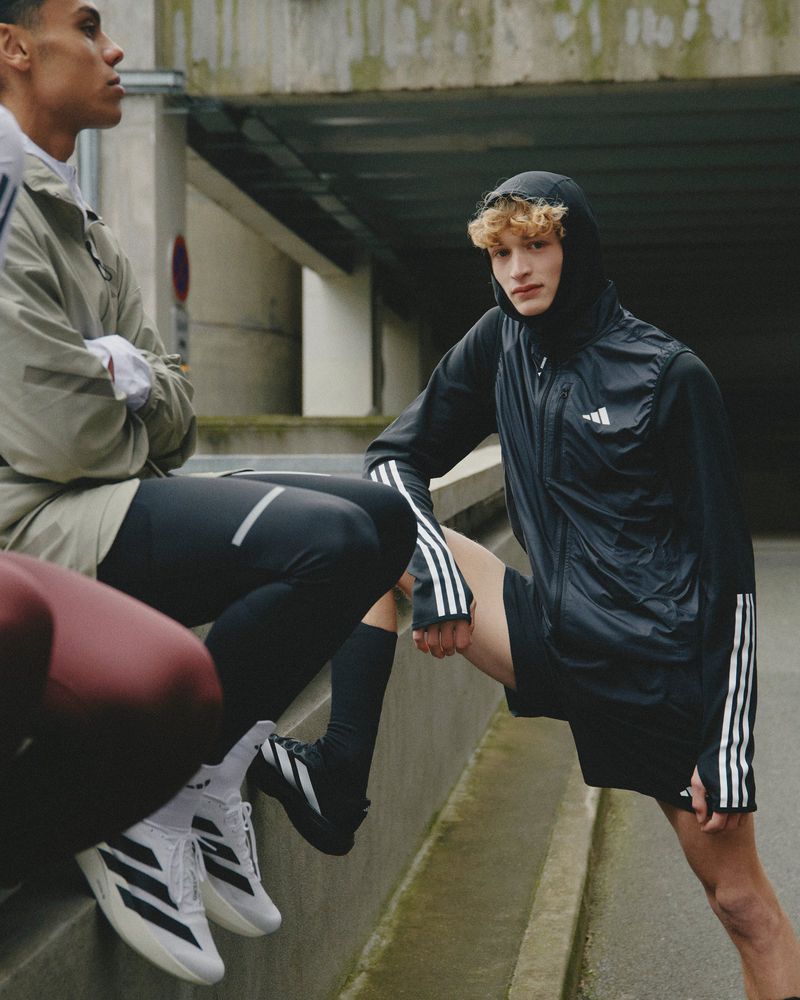 A group of male athletes are sitting and stretching outdoors in athletic apparel, in front of a concrete wall, wearing Adidas branded gear.