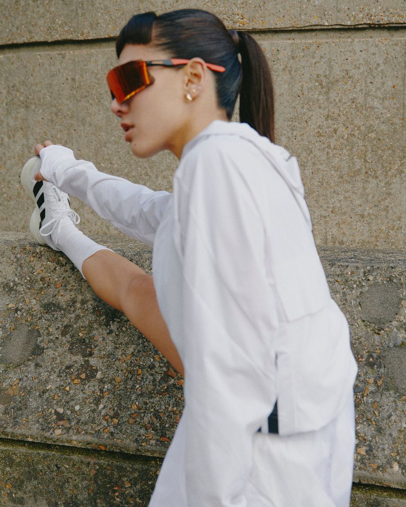 Woman in white sportswear stretches her leg in an urban setting, wearing orange sunglasses and white sneakers, posing against a concrete wall.