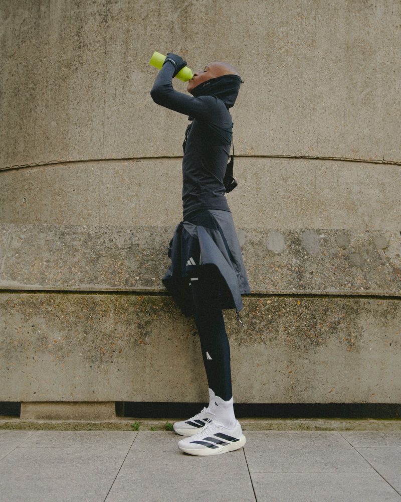 An athlete drinks water outdoors against a textured concrete background, wearing Adidas branded athletic wear.