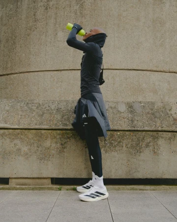 An athlete drinks water outdoors against a textured concrete background, wearing Adidas branded athletic wear.