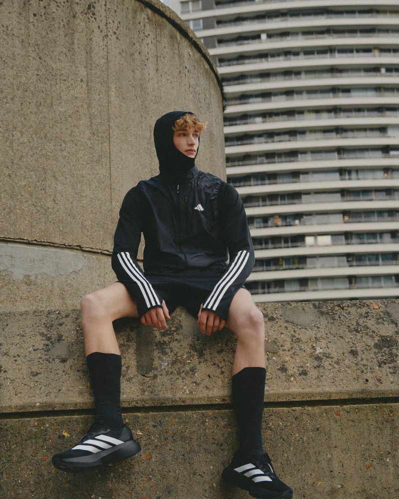 A young adult male sits on a concrete wall in athletic attire against a backdrop of an urban building. He wears a black jacket, shorts, socks, and sneakers.