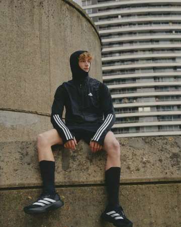 A young adult male sits on a concrete wall in athletic attire against a backdrop of an urban building. He wears a black jacket, shorts, socks, and sneakers.