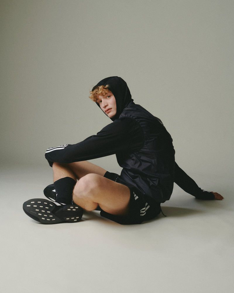 A young, fair-skinned man with curly blonde hair is sitting on the floor in a hooded track jacket, shorts, socks, and shoes, all Adidas.