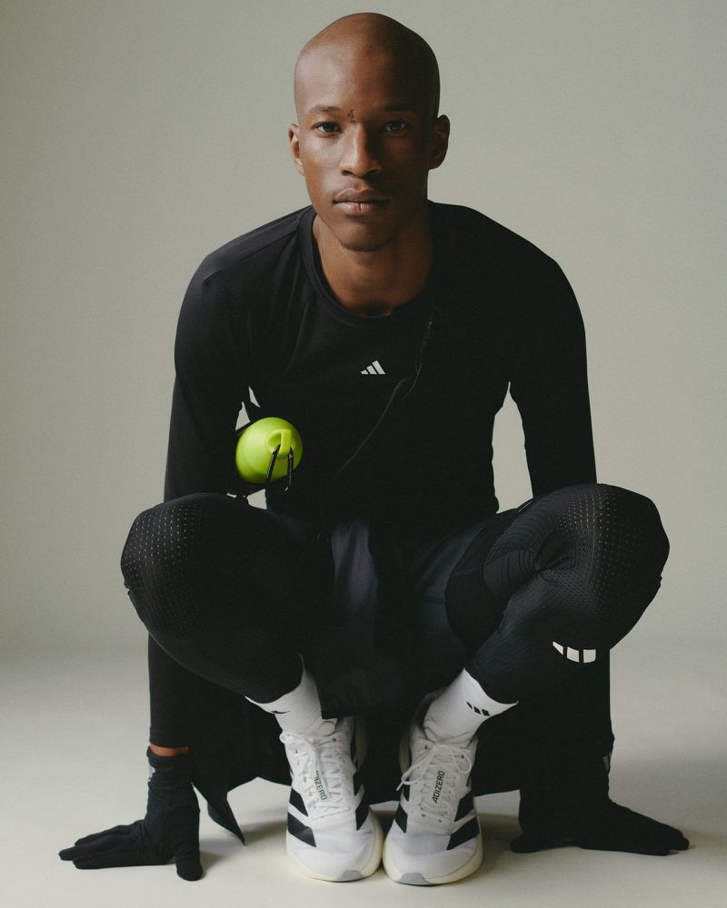 Athletic bald man in dark sportswear and white tennis shoes poses in a studio holding a yellow tennis ball. Fashion and sport