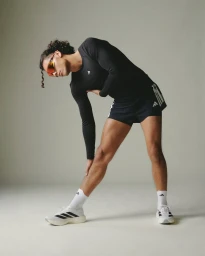 Male runner in black long-sleeve athletic shirt and shorts stretching his leg against a neutral studio backdrop.