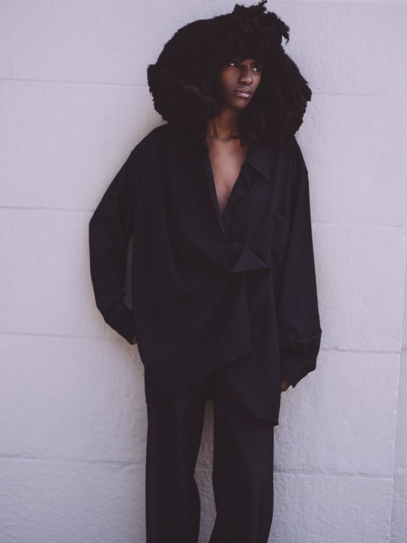 Editorial fashion shot of a Black adult female model with a large afro, wearing an oversized black button-down shirt and black pants against a white wall.