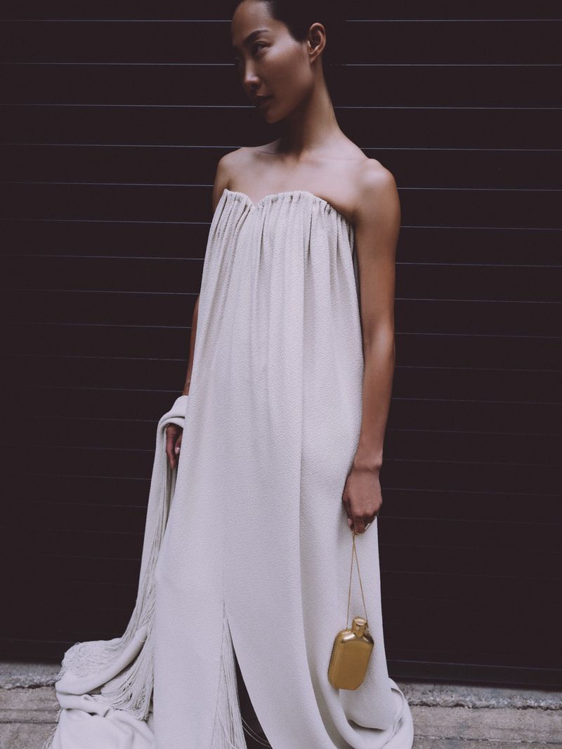Elegant woman wearing an off-white dress and holding a golden flask purse stands in front of a dark brown background.